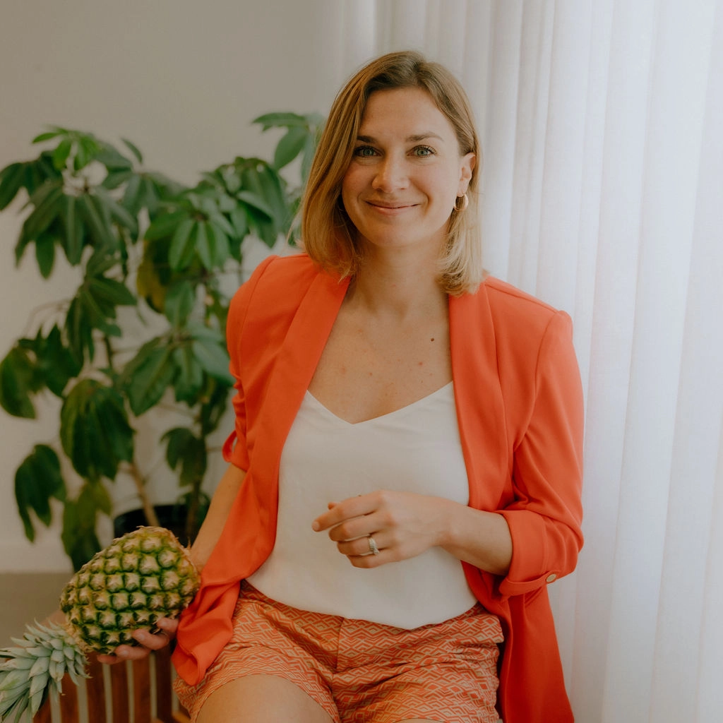 Femme souriante, assise sur une chaise, veston orange vif et chandail blanc. Elle tient un ananas dans ses mains