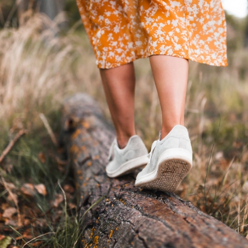 Femme en robe orange fleurie qui marche sur un tronc d'arbres couché au sol avec ses souliers blancs