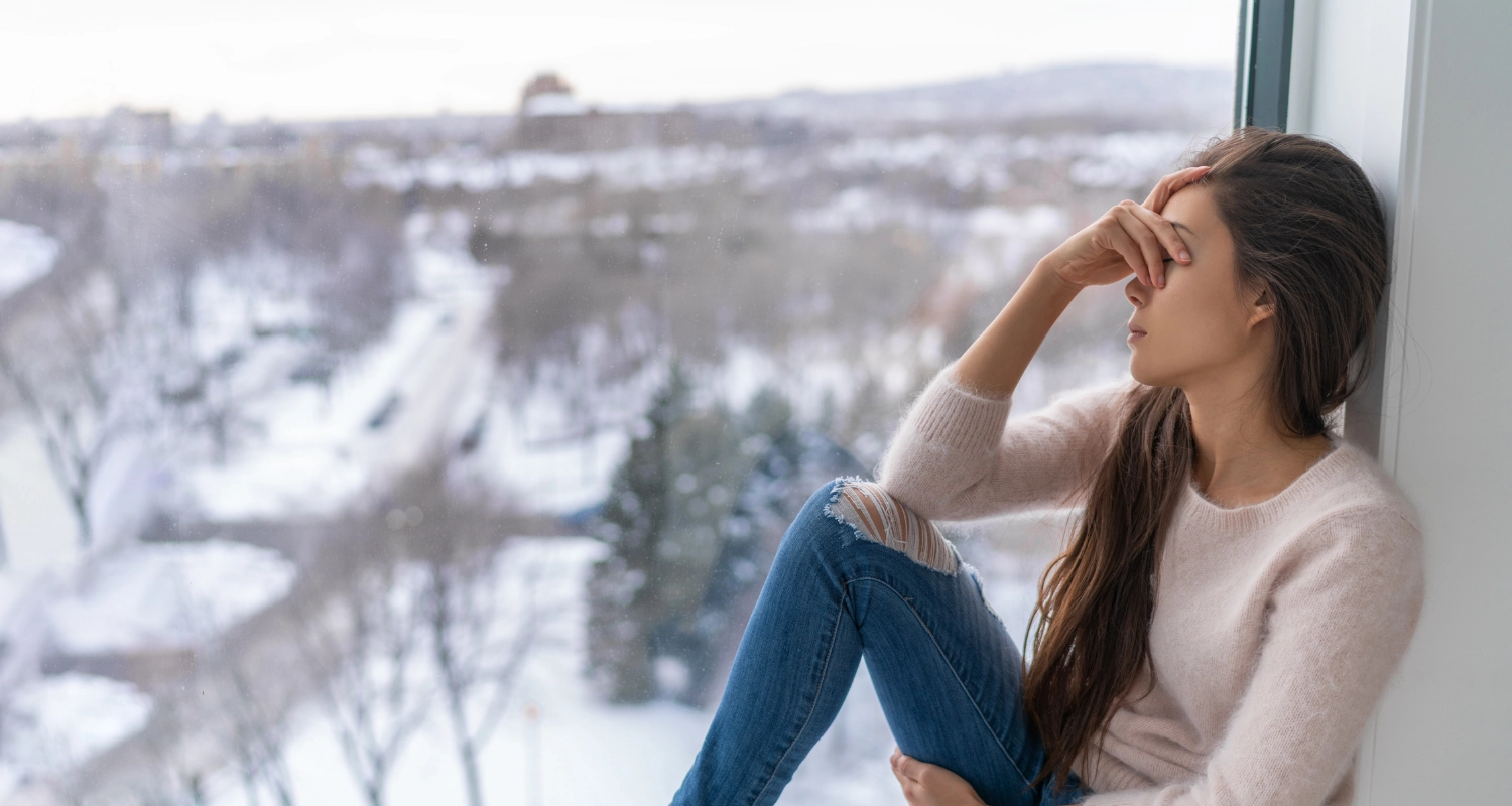 Femme assise sur le bord d'une fenêtre, paysage hivernal, mains qui cache ses yeux. Elle semble découragée ou fatiguée.