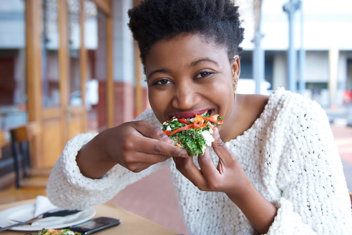 Femme souriante qui prend une bouchée d'une pizzafraiche avec de la roquette et des poivrons rouges