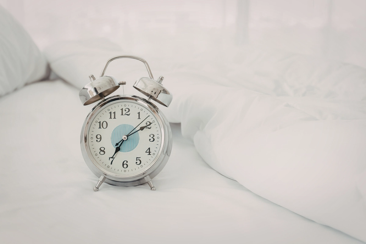 réveil matin argent avec centre bleu ciel, posé sur un lit couvert de draps blancs