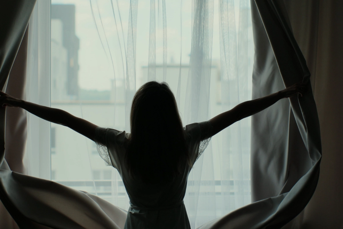 Silhouette de femme qui ouvre les rideaux en tissus épais devant une grande fenêtre avec un voile blanc