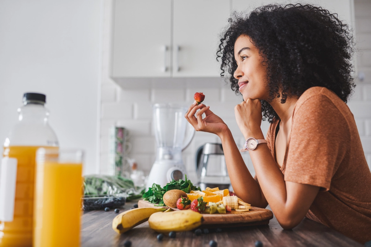 Femme, regard serain et sourire doux, qui tient une fraise dans ses mains. Devant elle se trouve divers fruits sur un comptoir